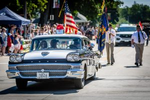Clear Lake, Iowa July 4 parade featuring a vintage police cruiser and VFW veterans carrying American flags down Main Avenue.